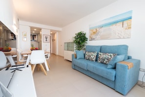Un salon moderne avec un canapé bleu, un coin repas avec des chaises blanches, un espace cuisine et un tableau sur le thème de la plage accroché au mur.