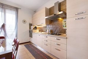 Eine moderne Küche mit Gasherd, Spüle und Edelstahlgeräten. Es gibt einen Essbereich mit Tisch und Stühlen, sowie ein Fenster mit durchsichtigen Vorhängen.