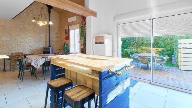 Una cocina con isla de madera, taburetes de bar, una mesa de comedor con sillas y vistas al jardín a través de grandes puertas correderas de vidrio.