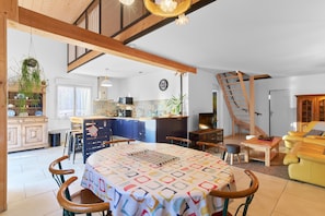 A spacious living area with a dining table, chairs, a sofa, a kitchen in the background, and a staircase. - Logement Pascal – Bright, Air-Conditioned House with Pool Table (Villeneuve-sur-Lot)
