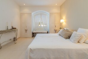Une chambre à coucher avec un grand lit, des tables de chevet, un banc et une fenêtre ornée de rideaux légers.