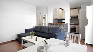 A modern living room with a grey sofa, a white coffee table, and a dining table with chairs. There is a kitchen area with wooden cabinets and a white refrigerator.