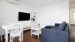 A living room with a white TV, a grey sofa, a white dining table with chairs, and a white cabinet.