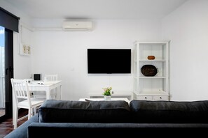 Uma sala de estar moderna com uma TV de tela plana, um rack branco para TV, um sofá, uma mesa de jantar com cadeiras e um pequeno armário branco.