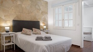 A bedroom with a stone wall, a bed with white bedding, two bedside tables with lamps, and a window.