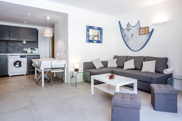 A modern living room with a grey sofa, white coffee table, and ottomans. There is a kitchen area with a washing machine and a dining table with chairs.