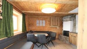 Ein gemütlicher Raum mit Holzbalkendecke, Steinwand, Kamin, Esstisch mit Stühlen und einer Couch.