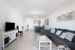 A modern living room with a flat-screen TV, a grey sofa, a white coffee table, and a dining area in the background.