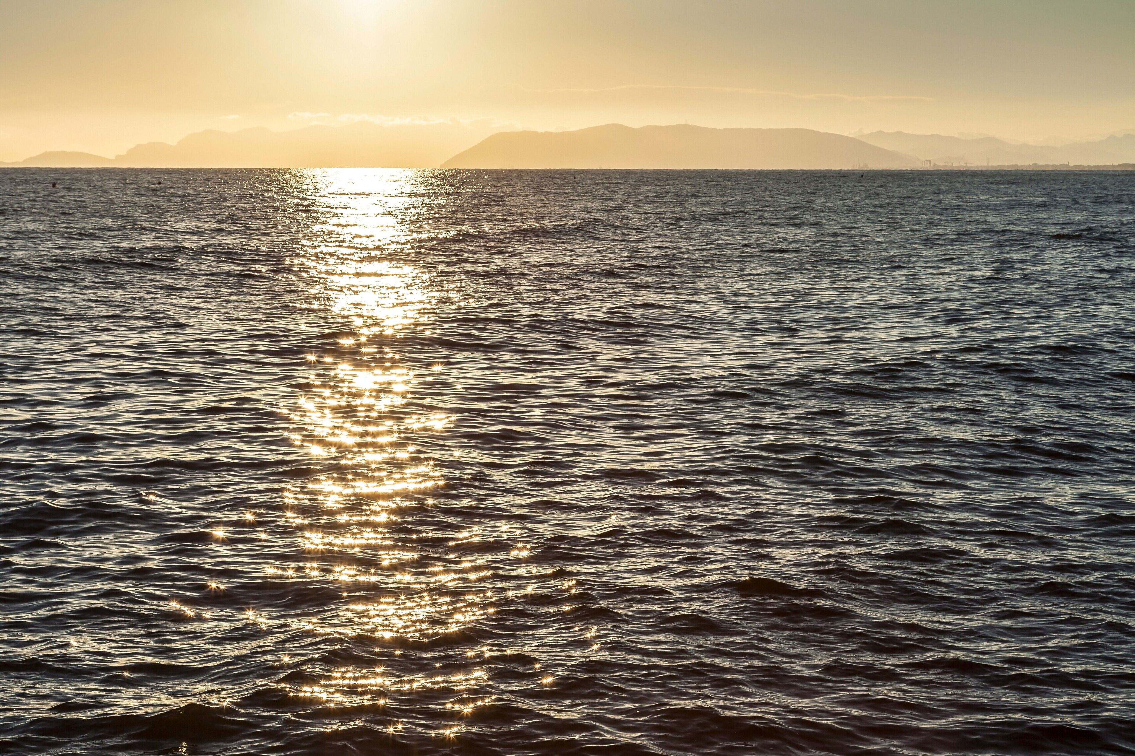 Une mer calme, avec la lumière du soleil se reflétant sur l’eau, des montagnes lointaines et un ciel dégagé.