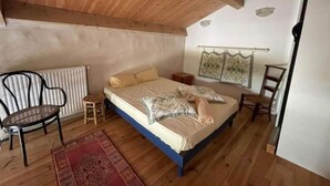 Een slaapkamer met een houten plafond, een bed met beddengoed met een patroon, een klein krukje, een stoel en een radiator.