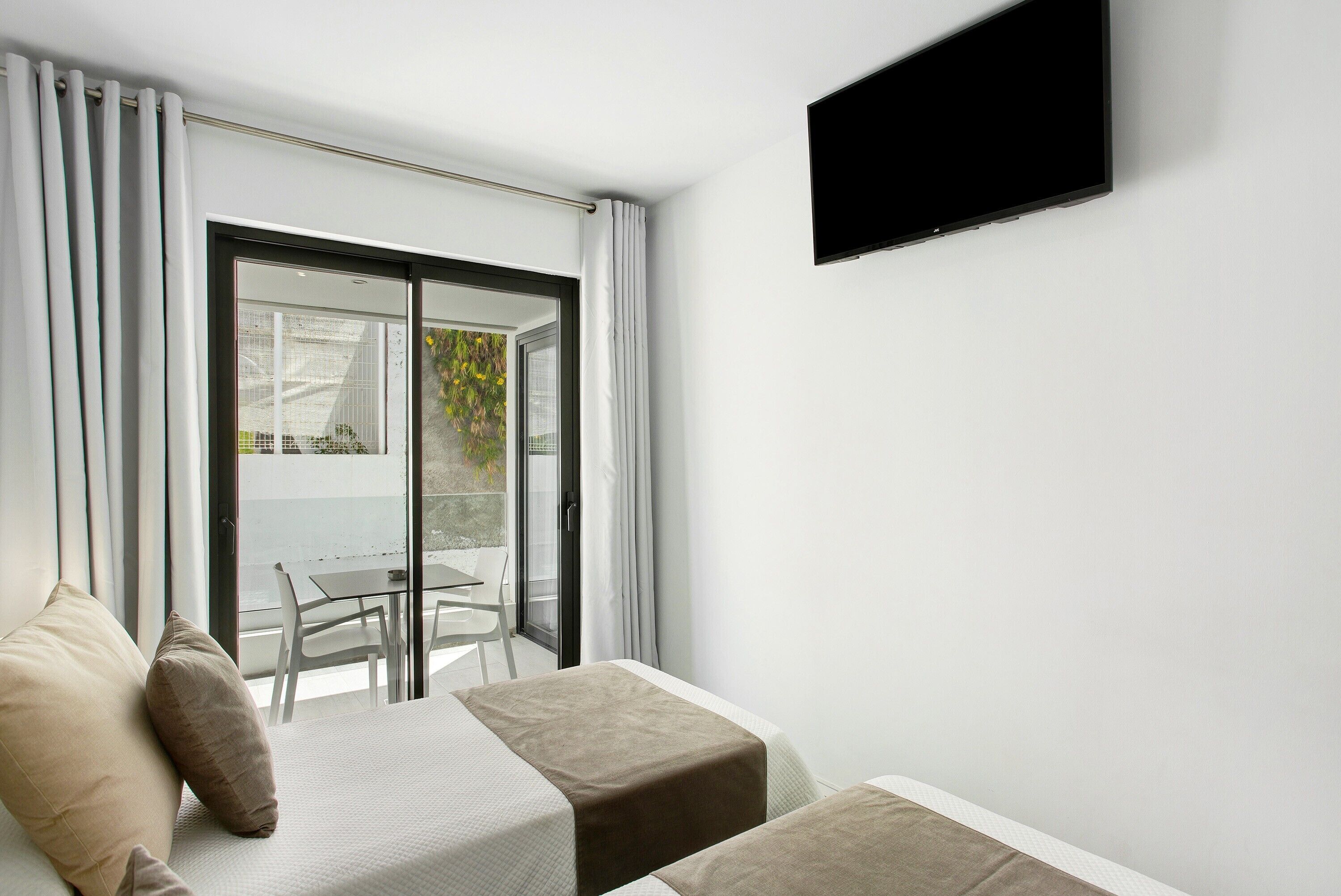 Una habitación moderna con una cama, un televisor y un ventanal corredera que da a un espacio exterior con una mesa y sillas.