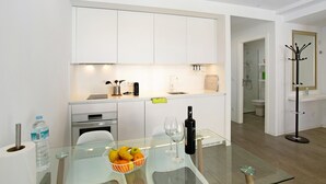 Une cuisine moderne avec des meubles blancs, une table en verre, un saladier rempli de fruits, des verres à vin et une bouteille de vin.