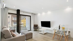 Un salon moderne comprenant un canapé, un pouf, une table basse en verre, une table à manger avec des chaises, un téléviseur à écran plat et un balcon offrant une vue sur les immeubles.