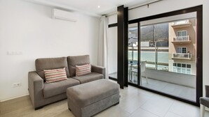Un salon moderne avec un canapé gris, un pouf assorti et une grande porte-fenêtre coulissante donnant sur un balcon avec vue sur les bâtiments et l’eau.