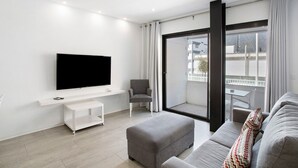 Ein modernes Wohnzimmer mit einem Flachbildfernseher, einem grauen Sofa, einem grauen Hocker, einer weißen TV-Konsole und einer Glastür, die auf einen Balkon führt.