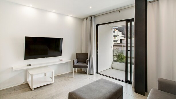 Una sala moderna con una televisión de pantalla plana, un sofá, una silla y un puerta corredera de cristal que da a un balcón exterior.