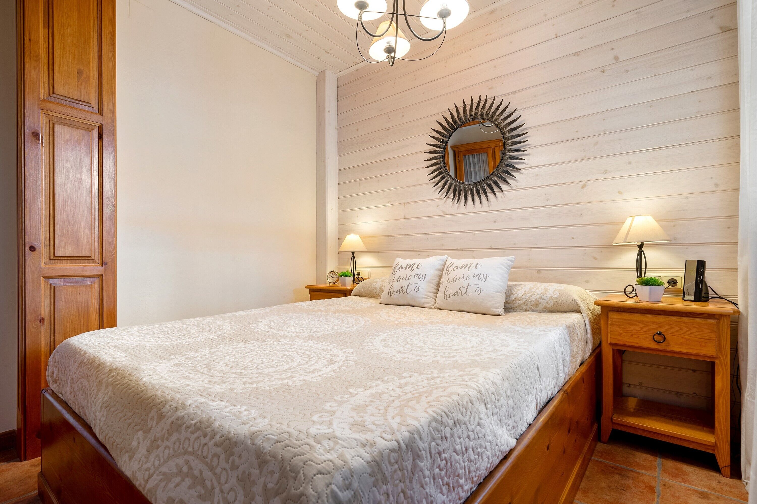 A bedroom with a large bed, wooden headboard, two bedside tables with lamps, a mirror, and a wall-mounted shelf.