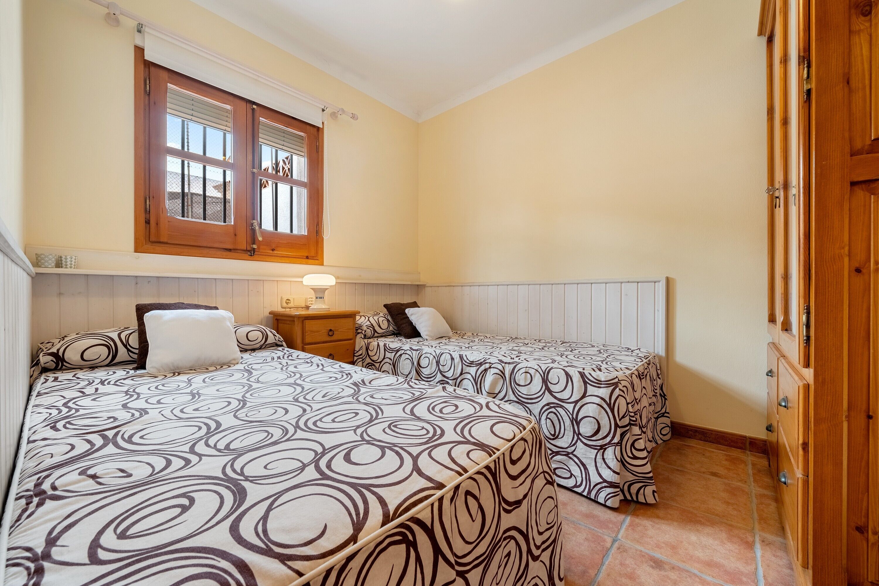 A bedroom with two beds, a window, a nightstand, and a wooden wardrobe.