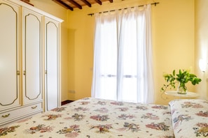 Un dormitorio con cama, armario, ventana con cortinas y una pequeña mesa con una planta.