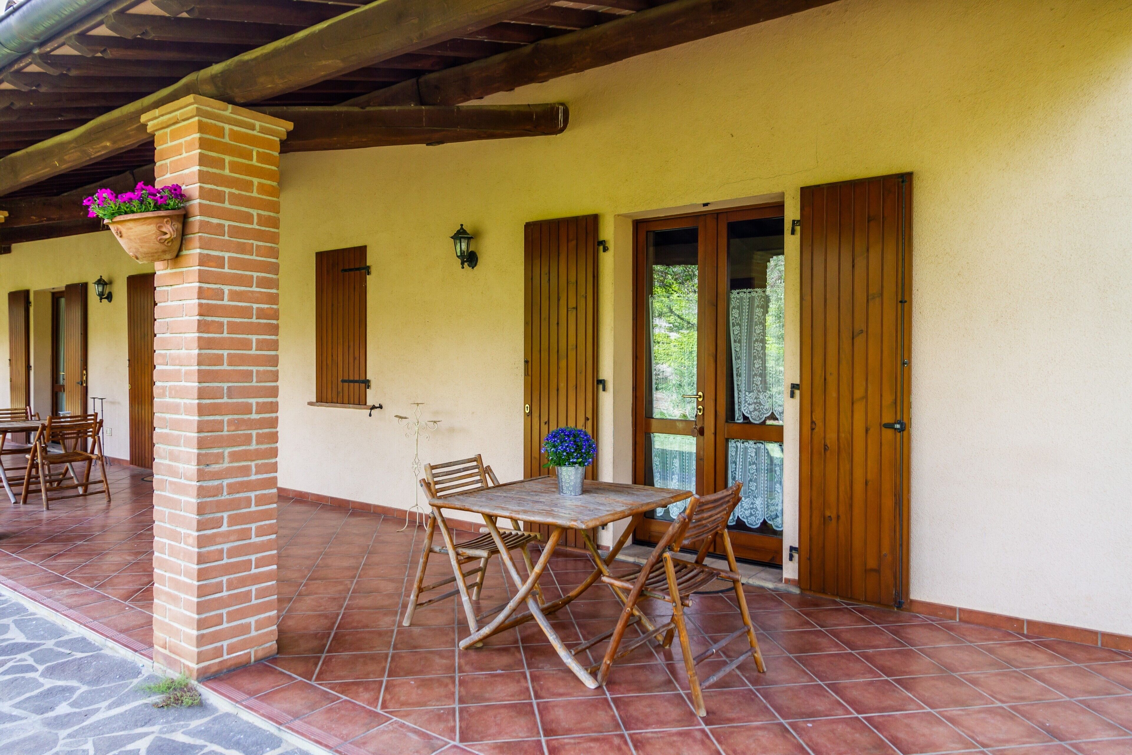 Eine überdachte Terrasse mit einer Ziegelstütze, einem Holztisch und -stühlen sowie einer Holztür mit Fenster.