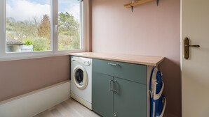 A laundry room with a washing machine, cabinets, a window, and a shelf with decorative items.