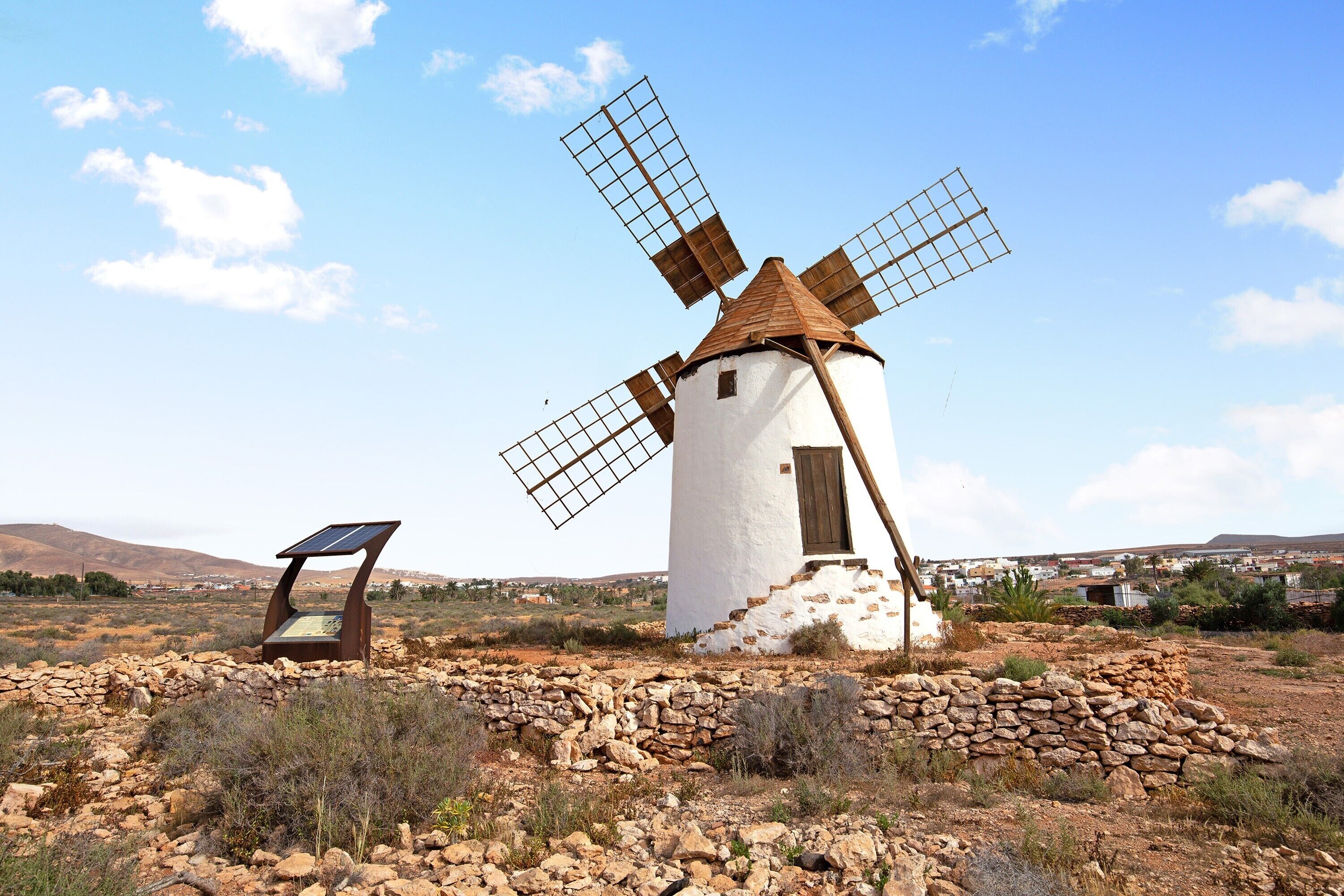 Un moulin à vent doté d’une base en pierre, d’une structure blanche et d’un toit brun, situé dans un paysage rural avec des collines en arrière-plan.