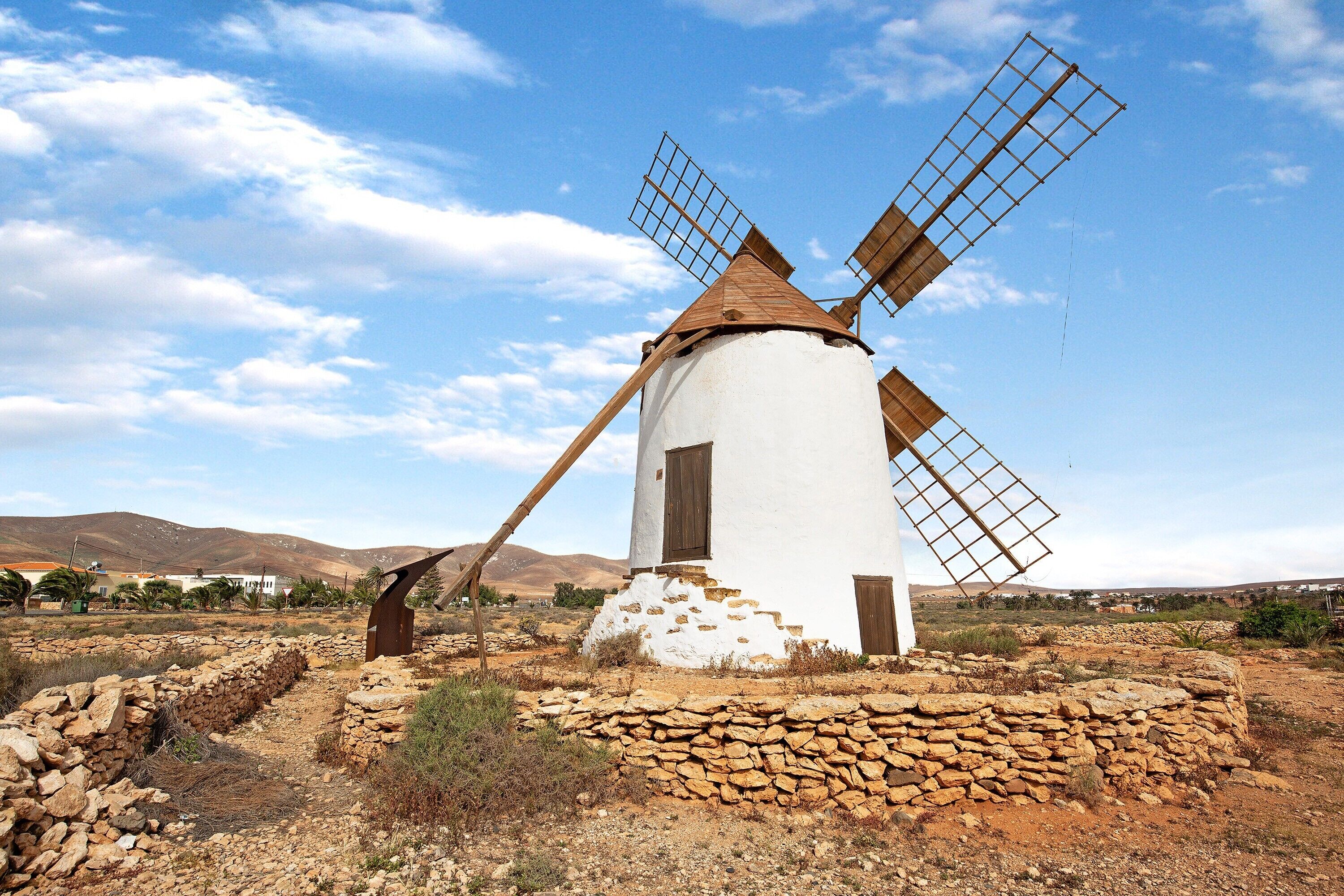 Un moulin à vent, avec un corps blanc et des pales en bois, se dresse dans un paysage aride, parsemé de murs de pierre et dominé par des collines lointaines.