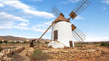 Un moulin à vent, avec un corps blanc et des pales en bois, se dresse dans un paysage aride, parsemé de murs de pierre et dominé par des collines lointaines.