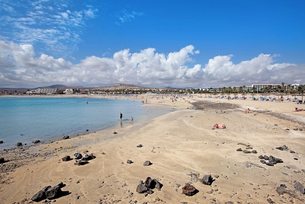A beach with clear water, rocky shore, and buildings in the background.