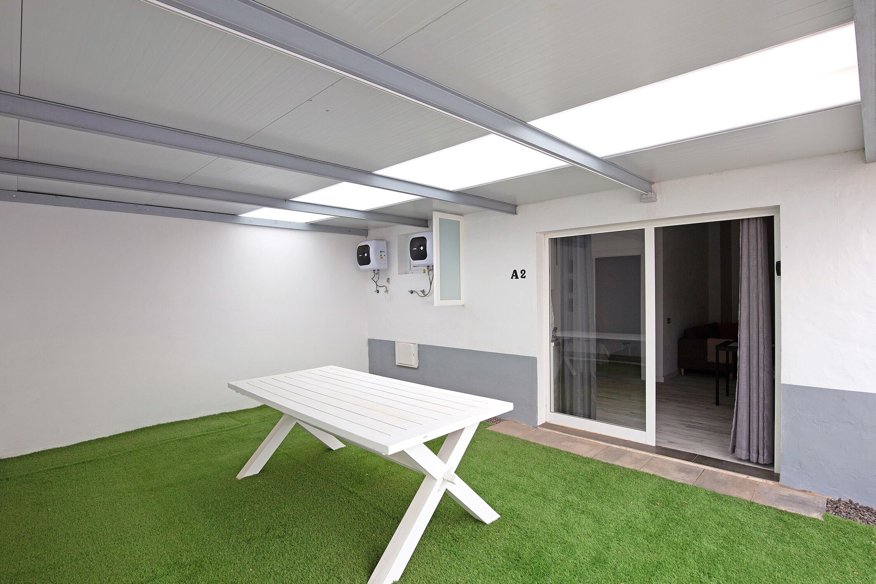 Ein Zimmer mit weißem Tisch, Kunstgrasanlage, Deckenleuchten und einer Schiebetür zu einem weiteren Raum.