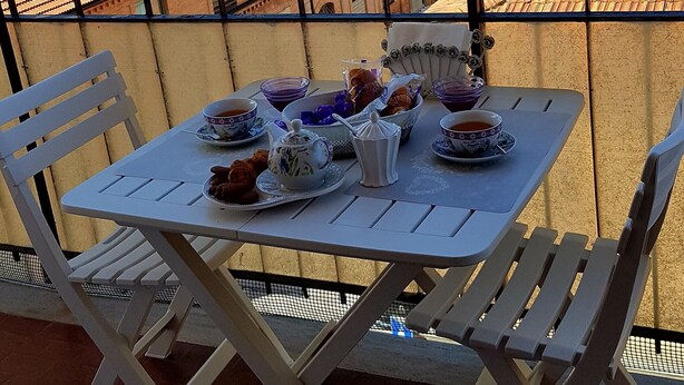 Een balkon met een tafel gedekt voor thee, stoelen en uitzicht op gebouwen en bomen.