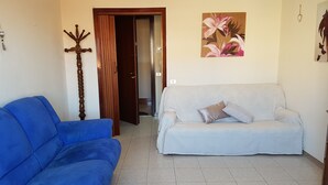 Een woonkamer met een blauwe bank, een lichtgekleurde bank, een houten kapstok en een bloemverf schilderij aan de muur.