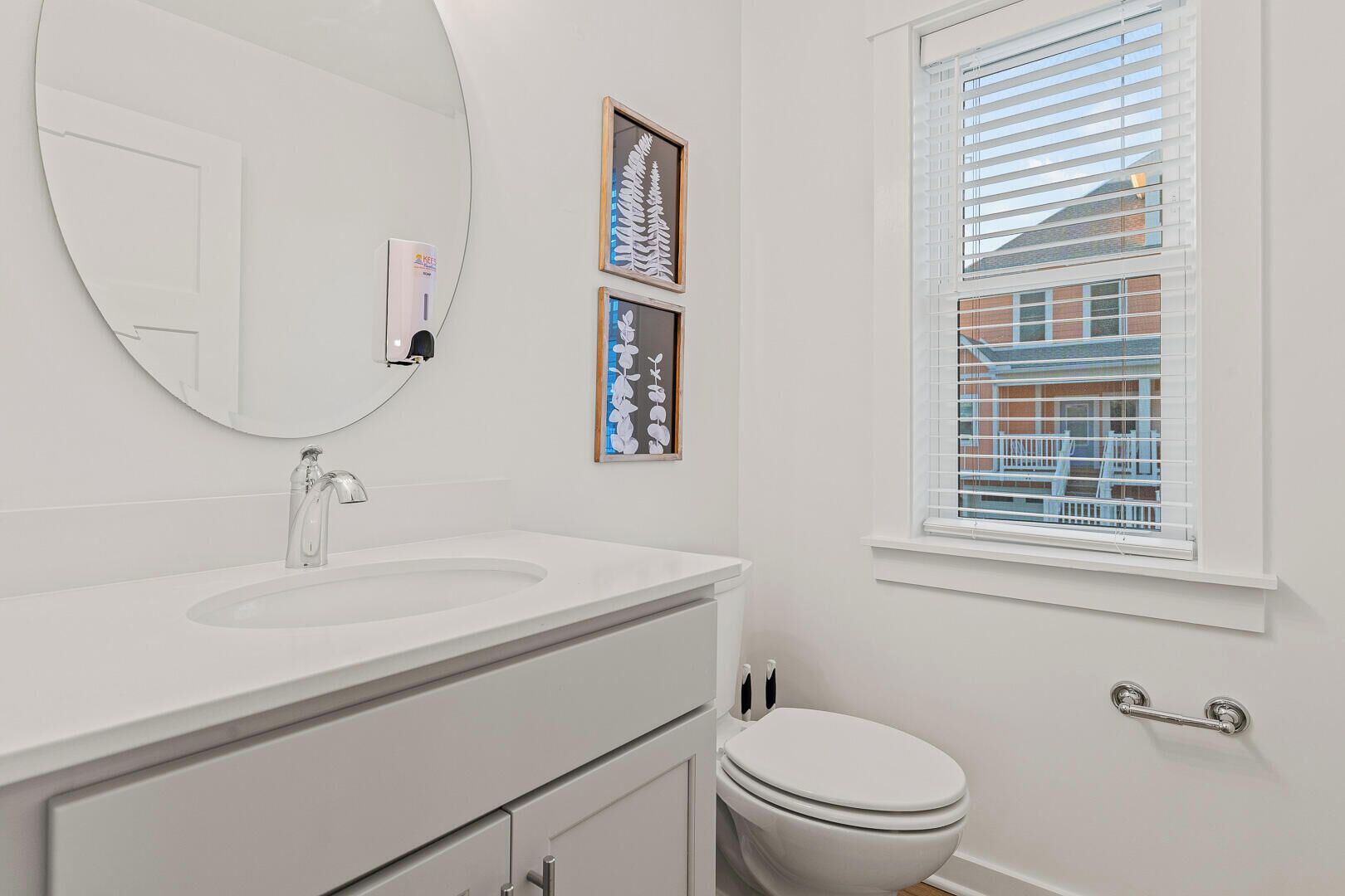 A bathroom with a white sink, a round mirror, a toilet, and a window with blinds.