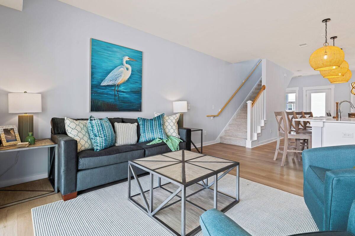 A modern living room with a gray sofa, patterned cushions, a glass coffee table, and a large painting of a bird. A staircase leads to a dining area with wooden chairs and a kitchen in the background.