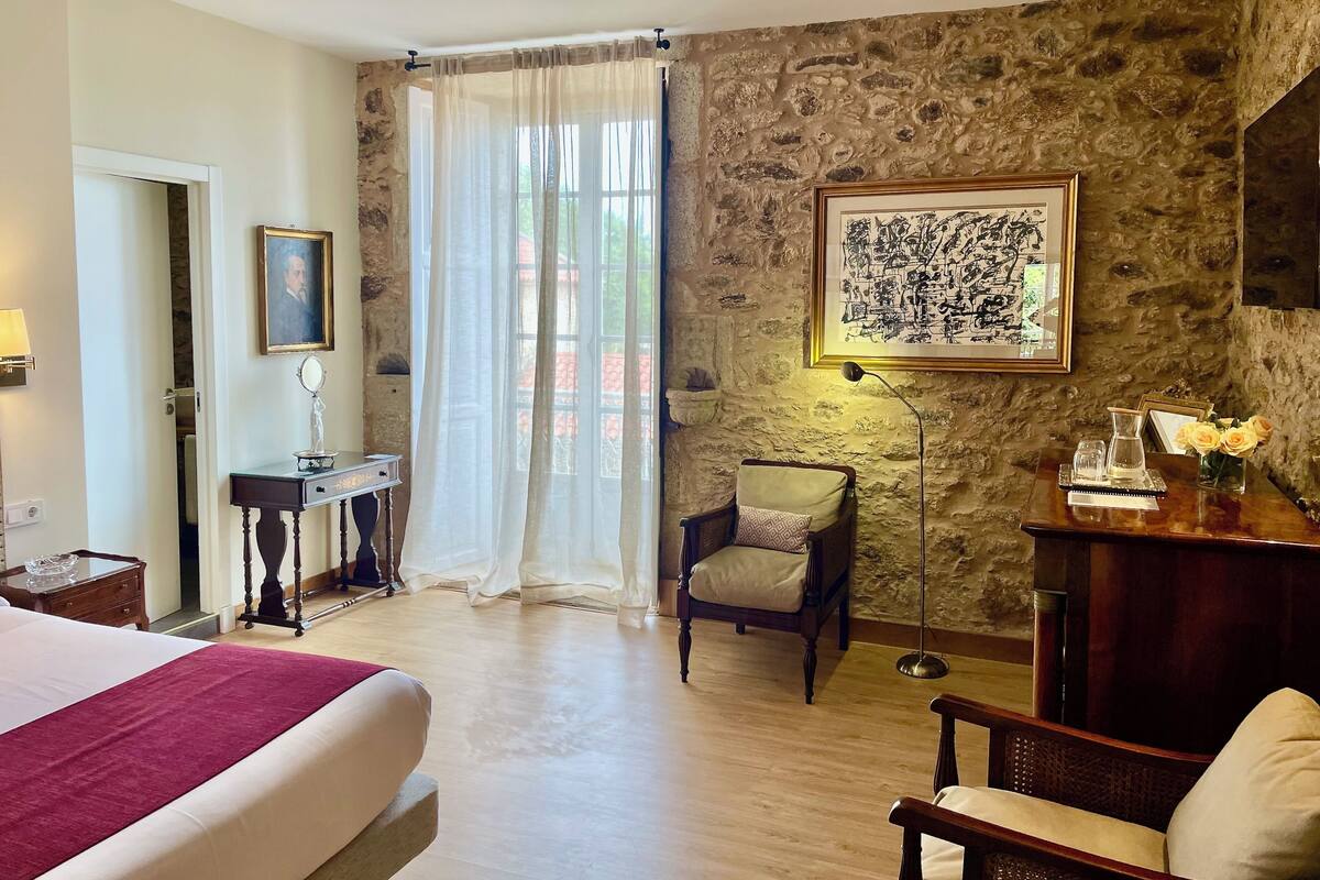Habitació d'hotel amb llit, una cadira, escriptori amb làmpada, quadre amb marc i paret de pedra.