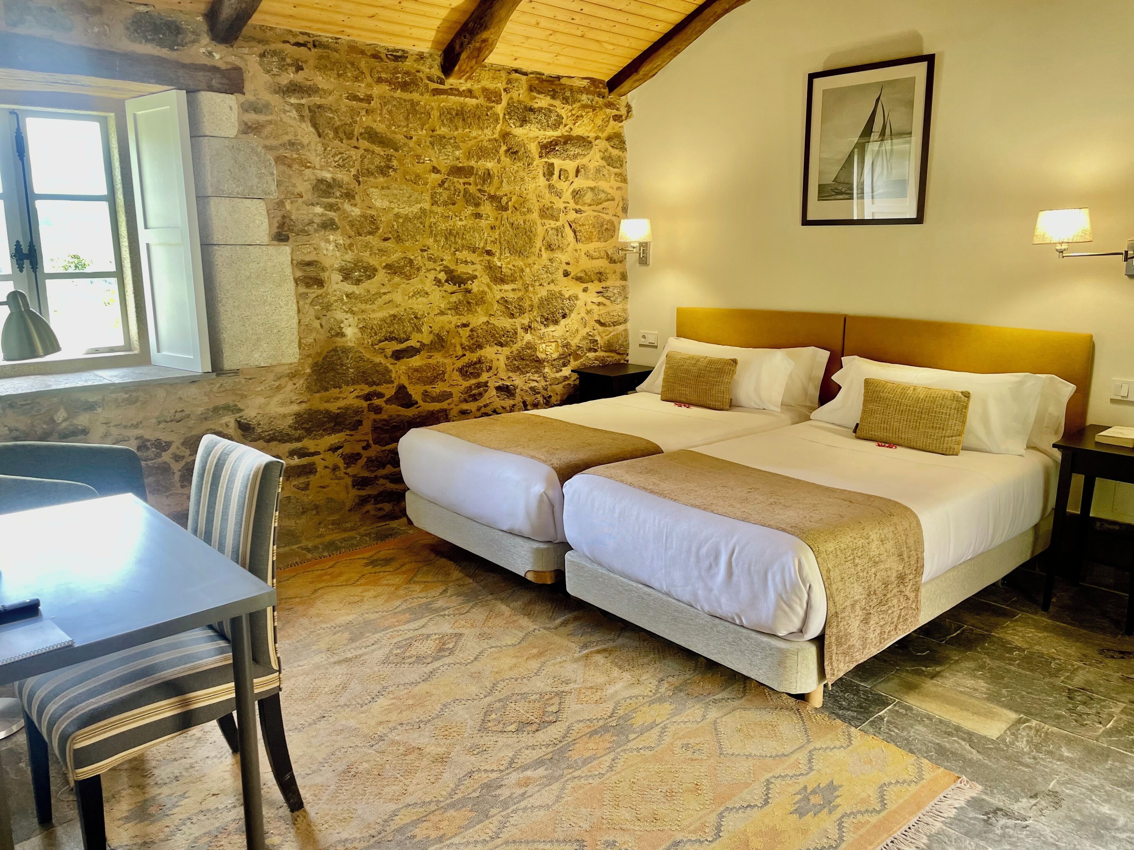 A double bed with a wooden headboard, a stone wall, a dining table with chairs, and a framed picture on the wall.