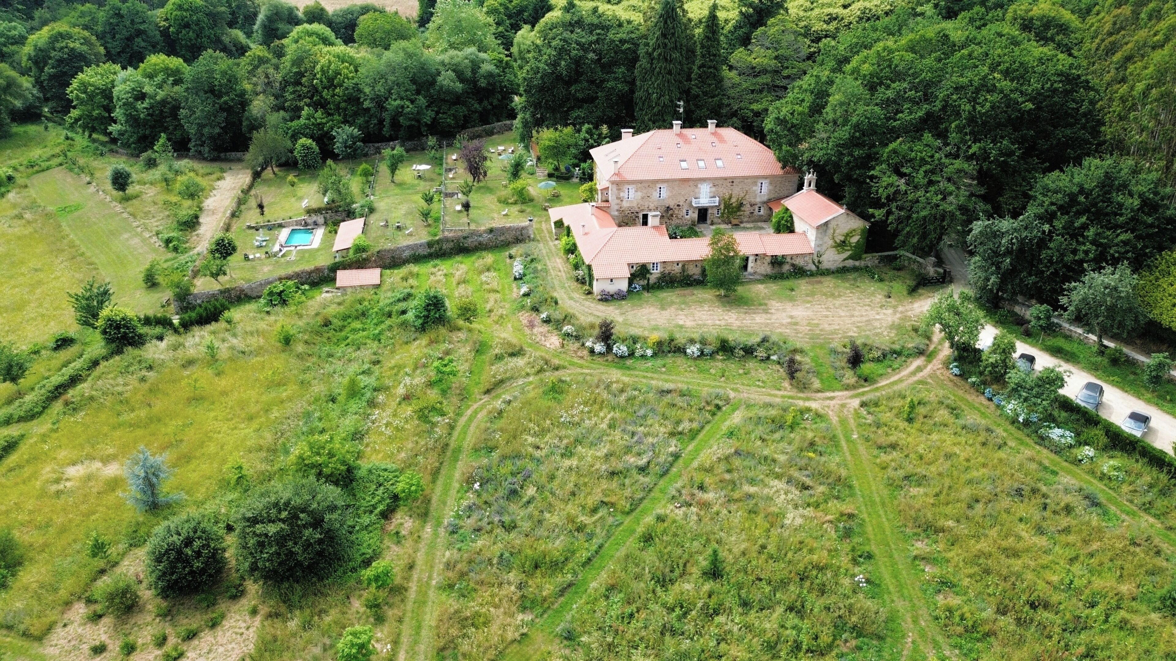 An aerial view of a large stone house with a red-tiled roof, surrounded by lush greenery and a swimming pool.