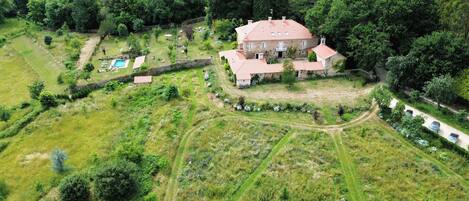 An aerial view of a large stone house with a red-tiled roof, surrounded by lush greenery and a swimming pool.