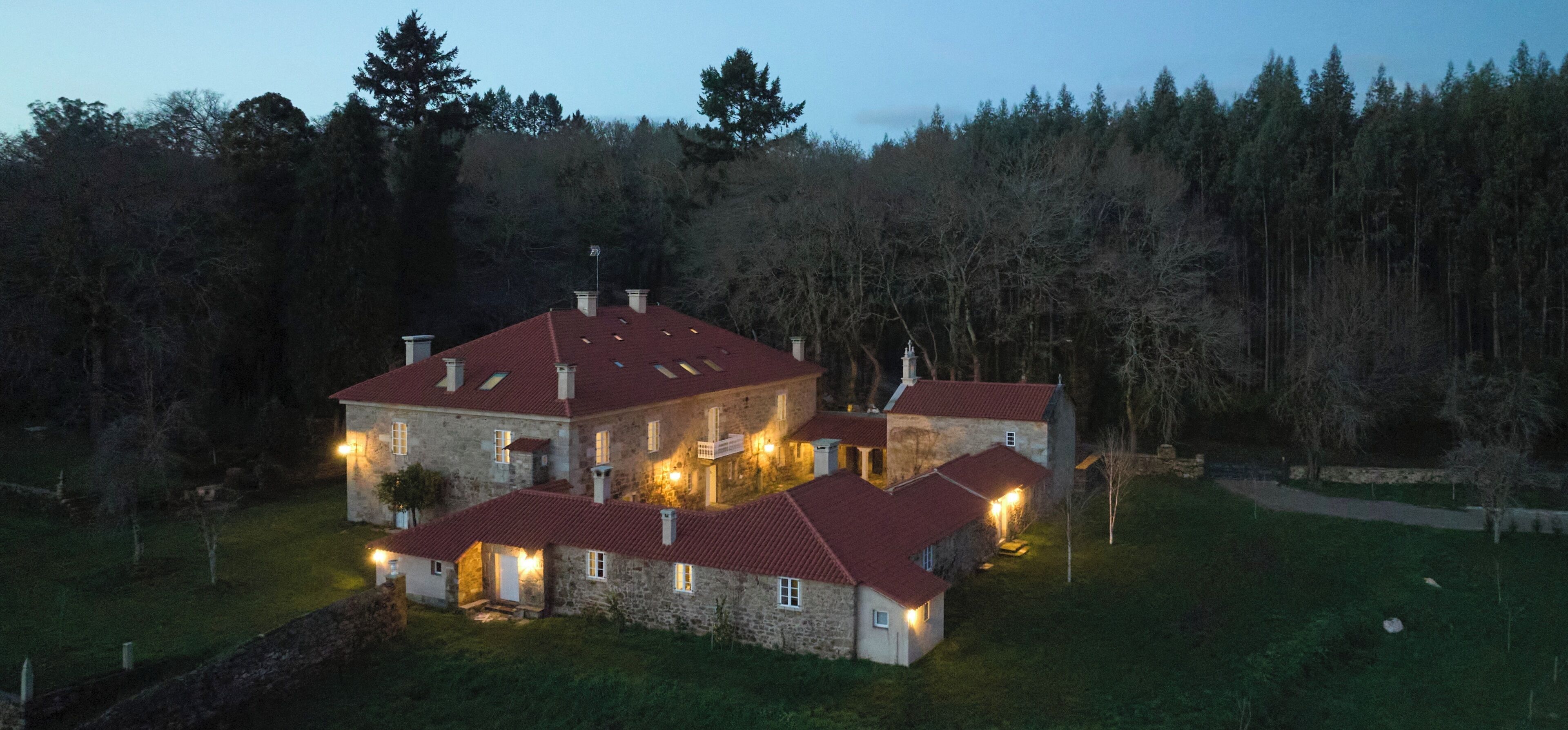 A large stone building with multiple red-roofed structures, surrounded by a forested area.