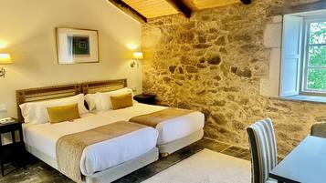 Una cama doble con cabecera de madera, dos mesitas de noche, un pequeño escritorio, una pared de piedra, una ventana con vistas y una mesa con una silla.