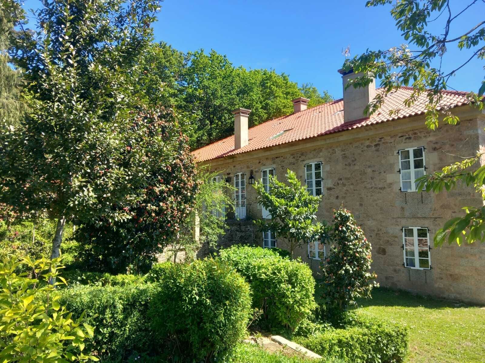 Un bâtiment en pierre, surmonté d’un toit recouvert de tuiles rouges, et entouré d’une végétation luxuriante et d’arbres.