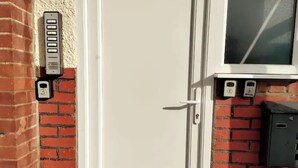 Une porte dotée d’un clavier pour l’ouverture, une boîte aux lettres et un coffret de raccordement électrique, le tout adossé à un mur de briques.