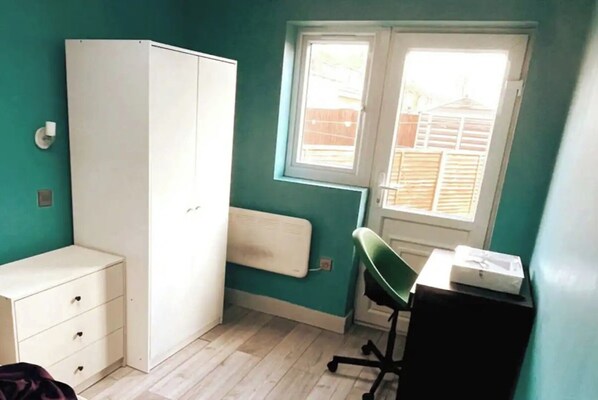 A small room with teal walls, a white wardrobe, a desk with a chair, and a window with blinds.