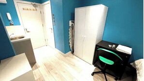 Une pièce avec un mur bleu, une armoire blanche, un bureau avec une chaise et une petite kitchenette.