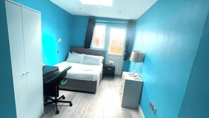 Une chambre à coucher de taille modeste, comprenant un lit, un bureau et une chaise. On y trouve une fenêtre avec des rideaux, une table de chevet et une lampe.