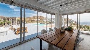 Ett rymligt rum med ett stort träbord, vita stolar och utsikt över en pool och ett kuperat landskap.