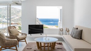 Ett modernt vardagsrum med ett stort fönster som vetter mot havet, och som inrymmer en soffa, fåtöljer och en tv.