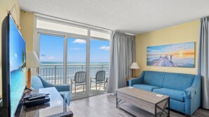 Une chambre d’hôtel avec un balcon, une télévision, un canapé, une table basse et un tableau représentant un coucher de soleil.