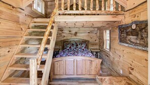 Interior de una cabaña de madera con un dormitorio en entrepiso, una escalera de madera y una cama con ropa de cama estampada.