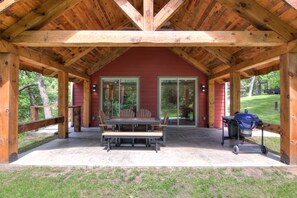 A covered patio with a wooden table and benches, a grill, and a red building with glass doors. - Amazing River House with covered patio! Sleeps 10-14. Kayaks,Fireplace,Games.  (Colfax)